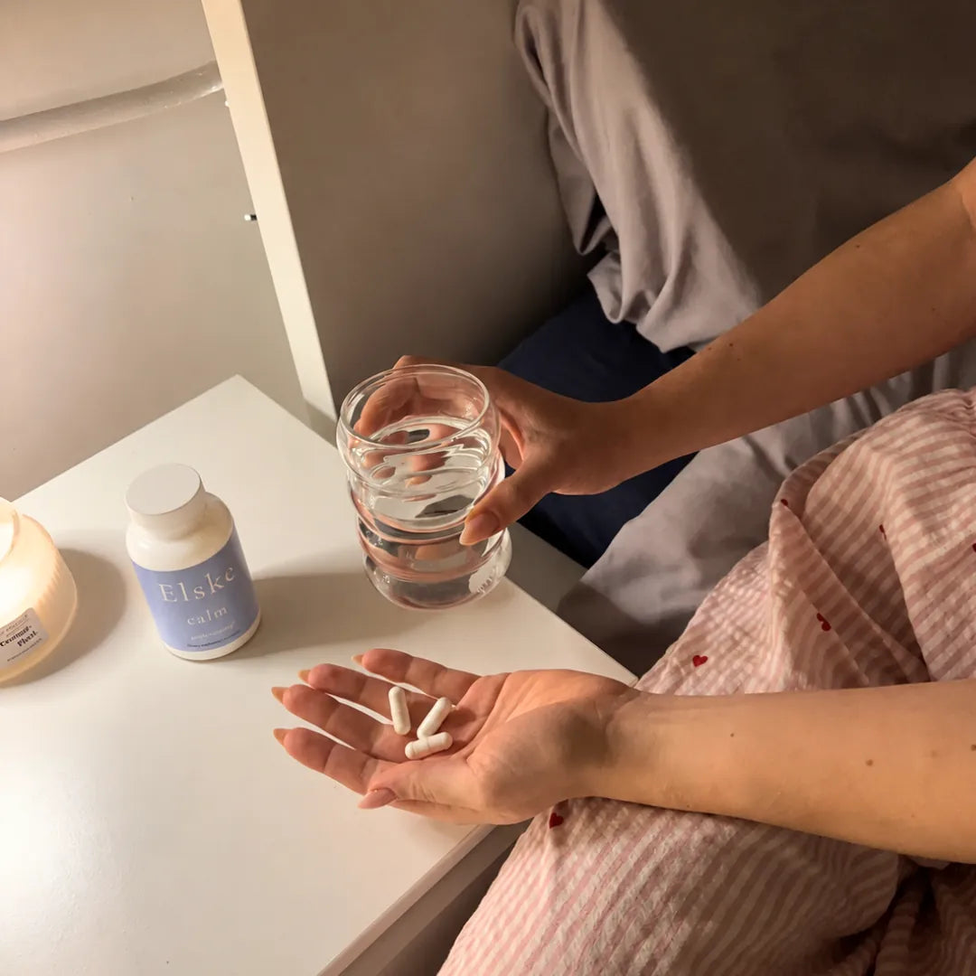 A person in pink striped pajamas holds a glass of water and three white capsules in their palm, with a bottle labeled 'Elske' on a white table nearby.